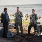 Drei Männer stehen an einem felsigen Strand, zwei in Uniformen mit einem Hund, einer in Lederjacke. Meer im Hintergrund.