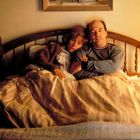 Ein Mann und eine Frau sitzen im Bett, umarmt unter einer gestreiften Decke, in einem gemütlich beleuchteten Raum.