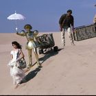 Vier Personen in der Wüste, eine Frau im weißen Kleid, ein goldener Roboter mit Sonnenschirm, Koffer werden getragen.