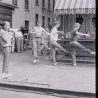 „MADELAINE“ und „HI-FI REPAIRS“ auf Schildern. Männer üben Tanzbewegungen auf der Straße.