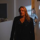 Zwei Frauen in einem schwach beleuchteten Raum; eine im Vordergrund in schwarzer Kleidung, die andere im Türrahmen in Silber.