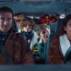 Ein Mann und eine Frau sitzen vorne im Auto, zwei Kinder hinten. Viele Geschenke sind im Hintergrund gestapelt.