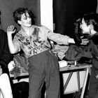 Drei fröhliche Personen interagieren ausgelassen, eine hält ein Getränk, eine Kamera ist sichtbar auf einem Tisch.