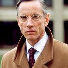 Ein Mann mit Brille und braunem Mantel, ernster Ausdruck, im Hintergrund unscharfe Gebäude.