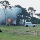 Ein brennendes Haus, umgeben von Bäumen. Ein Auto steht davor, eine Person sitzt auf einer Wiese und beobachtet.