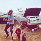 Zwei Männer neben einem Auto, aus dessen geöffnetem Kofferraum viele weiße Kaninchen herauslaufen. Einer der Männer ist gestürzt.