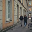 Zwei Männer gehen nebeneinander auf einem Bürgersteig entlang einer klassischen Fassade mit Bögen, rechts parken Fahrräder.