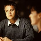 Ein Mann in einem grauen Pullover sitzt nachdenklich da, im Hintergrund eine Person unscharf.