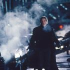 Eine Person in dunkler Kleidung geht durch eine neblige, nächtliche Stadtstraße voller Autos und Rauch.