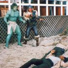 Zwei Figuren stehen am Strand: eine grün und muskulös, die andere in Rüstung. Im Vordergrund liegt eine Person mit "Sheriff" veste.