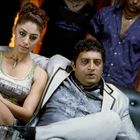 Eine Frau in einem Tanktop mit Text, ein Mann im Anzug, beide auf einem Sofa. Umgeben von lässig gekleideten Personen.