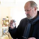 Ein Mann in einem dunklen Anzug und Schal untersucht konzentriert ein kleines Objekt. Im Hintergrund ein Bücherregal.