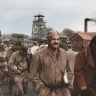 Eine Gruppe erschöpfter Arbeiter in schmutziger Kleidung, im Hintergrund ein Schachtgerüst und rauchender Schornstein.