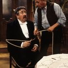 Zwei ältere Männer; einer in Anzug sitzt gefesselt auf einem Stuhl, der andere steht mit Seil in Arbeitskleidung.