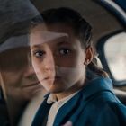 Ein Mädchen sitzt in einem Auto, schaut ernst aus dem Fenster. Das Gesicht einer älteren Frau spiegelt sich im Glas.