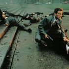 Ein Mann in einem Anzug kniet auf einem Bürgersteig mit vielen umgefallenen Personen herum. Er hält eine Waffe.