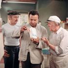 Drei Männer, einer hält eine Packung mit der Aufschrift "Farine spéciale PÂTISSERIE". Sie wirken konzentriert und diskutieren.