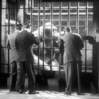 Drei Männer in Anzügen betrachten einen großen Affen hinter Gittern in einem schwarz-weißen Setting.