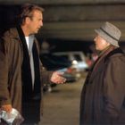 Zwei Personen unterhalten sich in einer Parkgarage. Links eine Person mit heller Jacke, rechts eine ältere Person mit Hut.