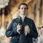 Ein Mann in aufwendiger historischer Kleidung, blickt ernst in einem eleganten Raum voller Menschen und Kronleuchter.