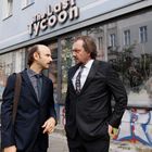 Auf einem Gehweg stehen zwei Männer in Anzügen vor einem Gebäude mit dem Schriftzug "The Last Tycoon".