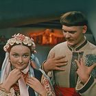 Ein Mann und eine Frau in traditioneller Kleidung; die Frau trägt einen Blumenkranz, im Hintergrund Holzmöbel.