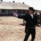 Ein Mann in schwarzer Kleidung zielt mit einem Revolver, im Hintergrund ein Haus und eine Gruppe von Kühen mit Cowboys.