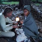 Ein Mann und eine Frau am Waldboden, bei Licht einer Laterne. Der Mann verbindet die Hand der Frau.