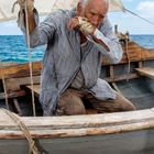 Ein älterer Mann in zerschlissener Kleidung sitzt auf einem Holzboot, das vor einem blauen Meer segelt.