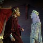 Ein König und eine Königin in prächtigen Gewändern stehen sich gegenüber. Der Raum hat rote Vorhänge und ein Bett.