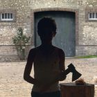 Ein Junge im Schatten hält eine Axt vor einem steinernen Gebäude mit Hühnern im Hintergrund.