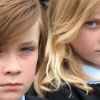 Zwei Kinder mit ernsten Gesichtsausdrücken sitzen auf einer Bank. Beide tragen Schuluniformen.