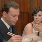 Ein Mann und eine Frau essen mit Essstäbchen in einem Restaurant mit Holzvertäfelung. Sie unterhalten sich.