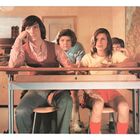 Der Tisch zeigt die Nummer "4". Vier Jugendliche sitzen gelangweilt in einem Klassenraum, Landkarte und Stadtbild im Hintergrund.