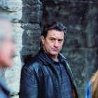 Ein Mann in einer schwarzen Lederjacke steht skeptisch an einer Steinwand. Eine andere Person ist im Vordergrund unscharf.