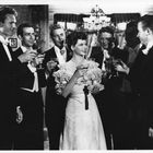 Personengruppe in festlicher Kleidung stößt mit Gläsern an, lächelnd in einem eleganten Raum mit Kronleuchter und Deko.