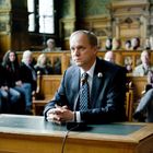 Ein Mann in einem Anzug sitzt an einem Tisch in einem Gerichtssaal. Hinter ihm sitzen Menschen auf Sitzbänken, die zuschauen.