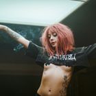 Frau mit roten Haaren und Tattoos tanzt unter hellem Licht, trägt ein schwarzes T-Shirt mit weißem Schriftzug.
