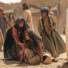 Eine Gruppe von Menschen in traditioneller Kleidung in einer Wüstenumgebung. Eine Person kniet am Boden.
