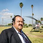 Ein Mann im Anzug steht auf einer Wiese mit Palmen; ein Hubschrauber fliegt im Hintergrund.