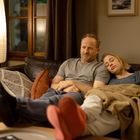 Ein Mann und eine Frau sitzen auf einem Sofa. Die Frau lehnt sich an den Mann und schläft. Eine Lampe strahlt warmes Licht.