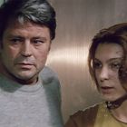 Ein Mann und eine Frau schauen aufmerksam durch ein Fenster mit Regentropfen. Er trägt grau, sie hat braune Haare.