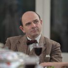 Ein Mann in einem braunen Anzug mit Fliege sitzt am Tisch. Vor ihm steht ein Glas Rotwein.