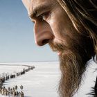 Ein Mann mit langem Haar und Bart schaut auf eine lange Karawane, die sich durch eine weiße Winterlandschaft schlängelt.
