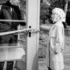 Ein Mann im Anzug in einer Telefonzelle, deren Tür mit einem Seil zugebunden ist, lächelt eine Frau mit Kopftuch an.