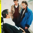 2001040445454SWR_F03. Zwei Personen stehen, eine im blauen Mantel, und sprechen mit einer sitzenden Person im Anzug.