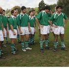 Sportler in grünen Trikots hören einem Mann mit Rugbyball zu, stehen auf einem Rasenfeld.