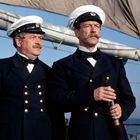 Zwei Männer in Marineuniformen mit Mützen stehen auf einem Segelschiff. Einer hält ein Fernglas. Der Himmel ist klar.