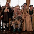 Mehrere Menschen in prunkvollen Barockkostümen stehen in einem opulent dekorierten Raum.