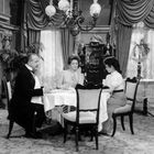 Drei Personen in eleganter Kleidung sitzen an einem Tisch in einem luxuriösen, historisch eingerichteten Raum.